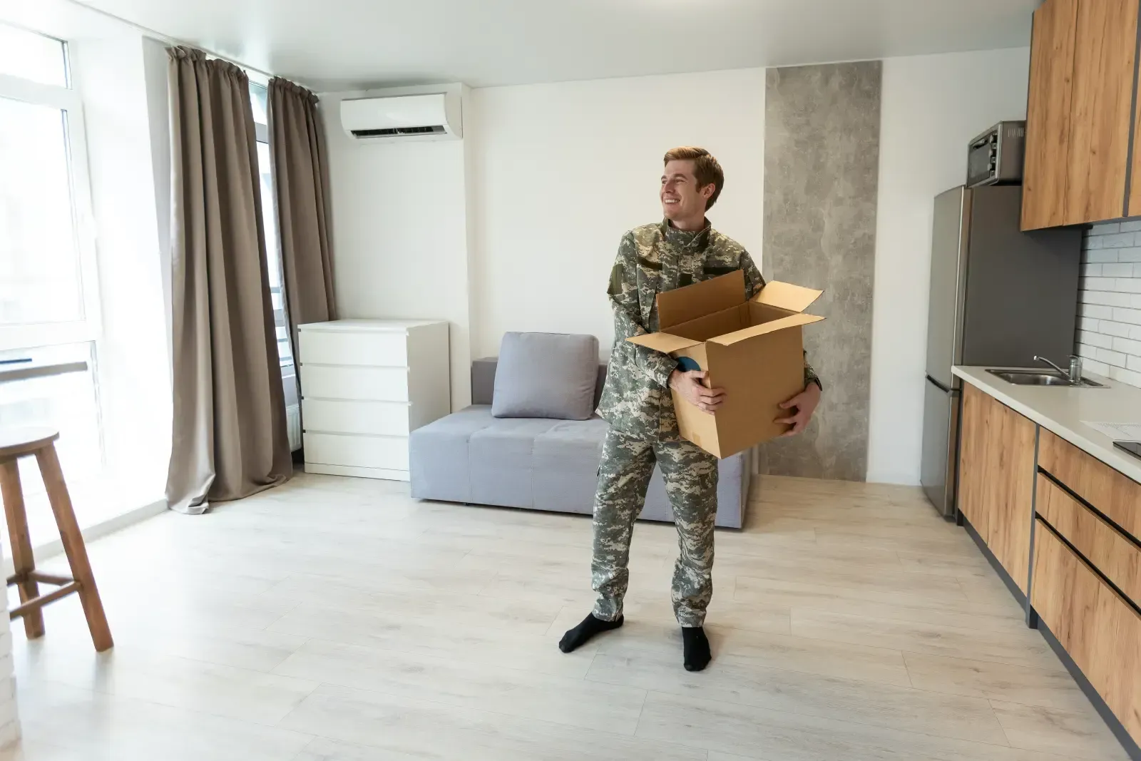 Man in camouflage uniform holding a box in a new apartment with an open kitchen.
