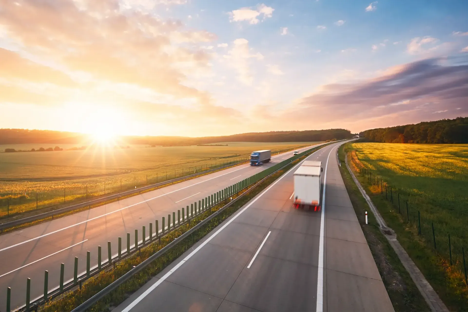 Trucks driving on a highway at sunset, with a field on the right and trees in the background.