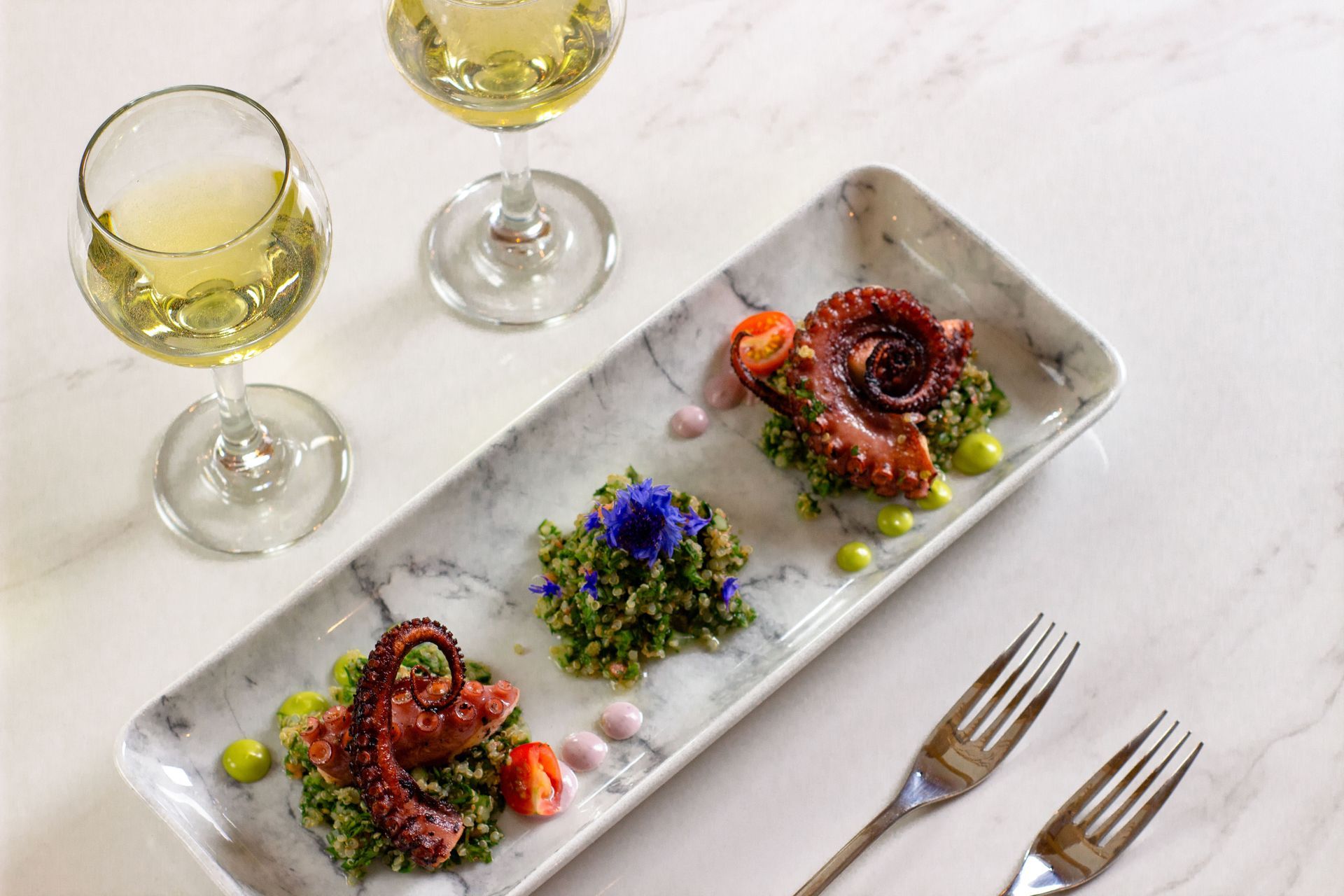 Elegante aperitivo de pulpo emplatado con microvegetales y flores comestibles, acompañado de dos copas de vino blanco.