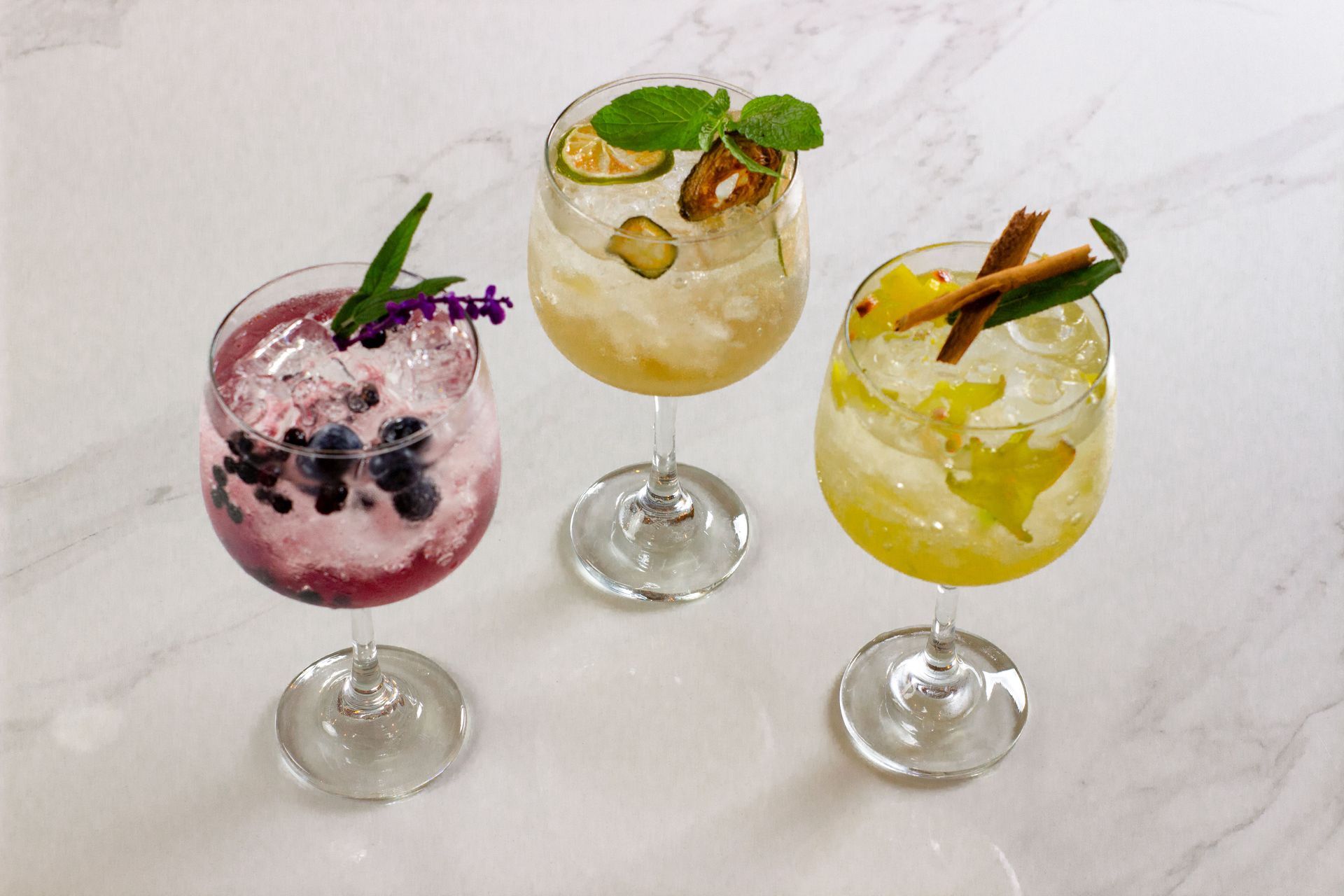 Tres coloridos cócteles helados en copas con tallo sobre una superficie blanca, adornados con menta y fruta.