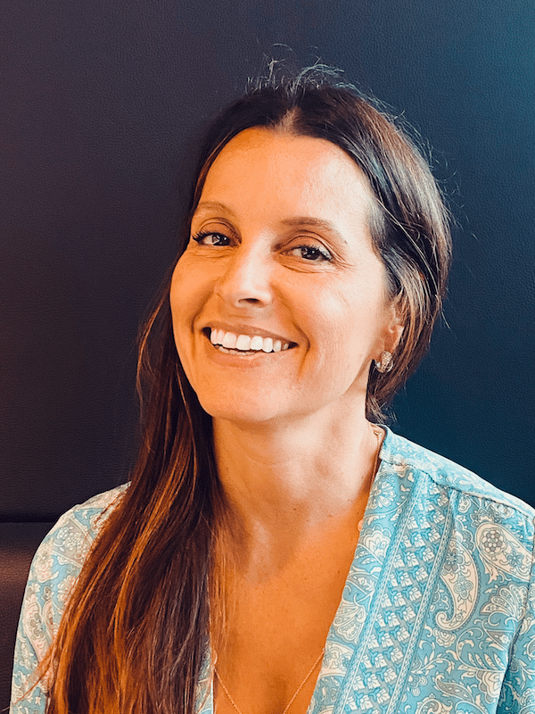 Femme souriante, vêtue d'un haut bleu à motifs, sur fond sombre.