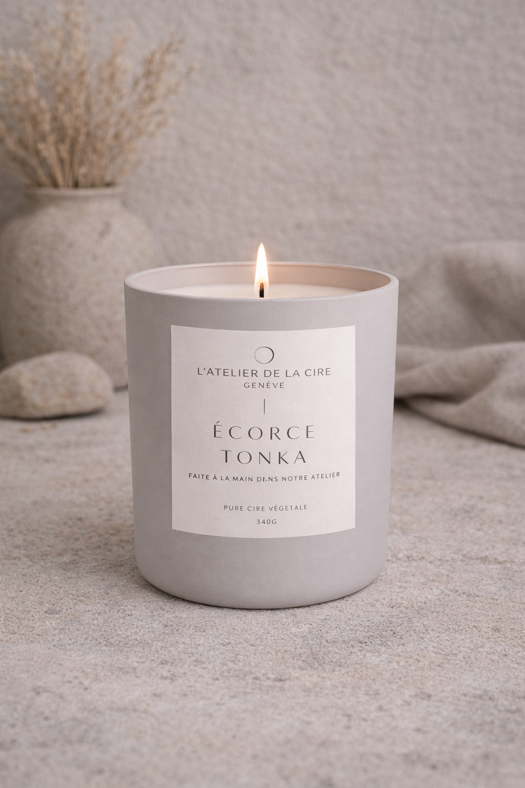 Bougie blanche allumée dans un pot en béton minimaliste, posée sur une surface grise avec des fleurs séchées en arrière-plan.