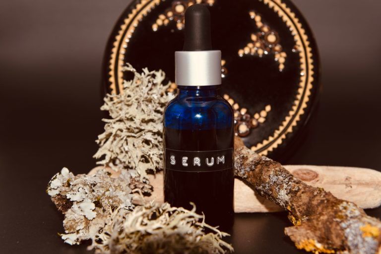 Flacon de sérum bleu avec bouchon argenté sur des fleurs séchées, le tout sur un fond décoratif sombre.