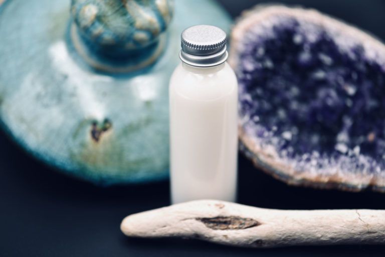 Petite bouteille blanche à bouchon argenté posée sur un morceau de bois flotté, à côté de verre de mer bleu et violet.