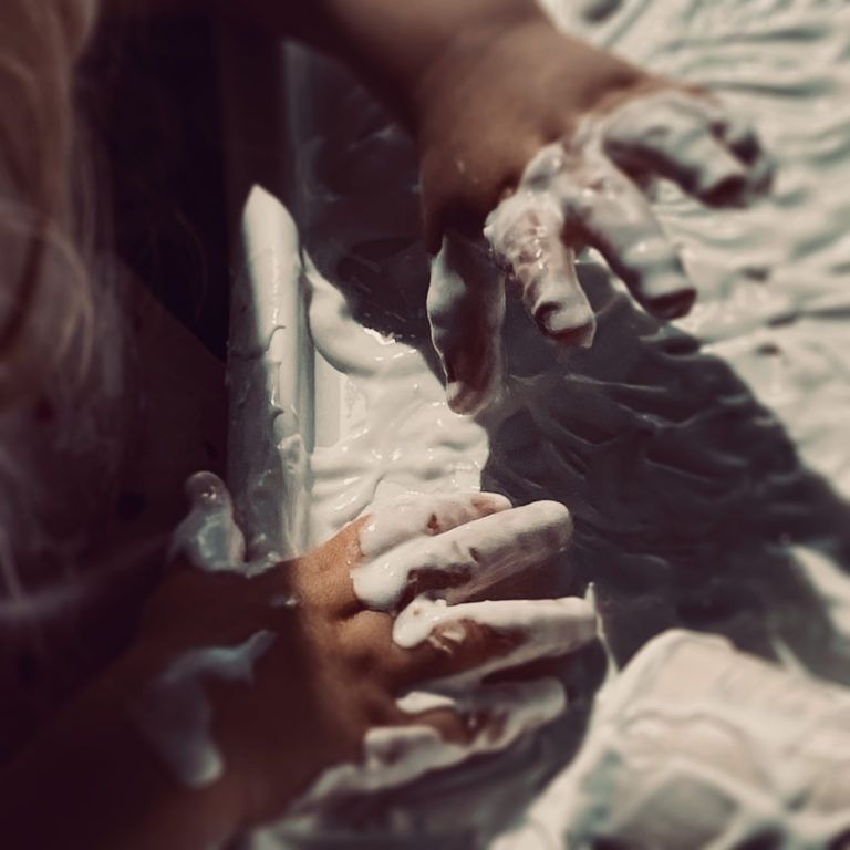 Des mains couvertes de crème blanche pétrissent une masse molle dans une lumière chaude et tamisée.