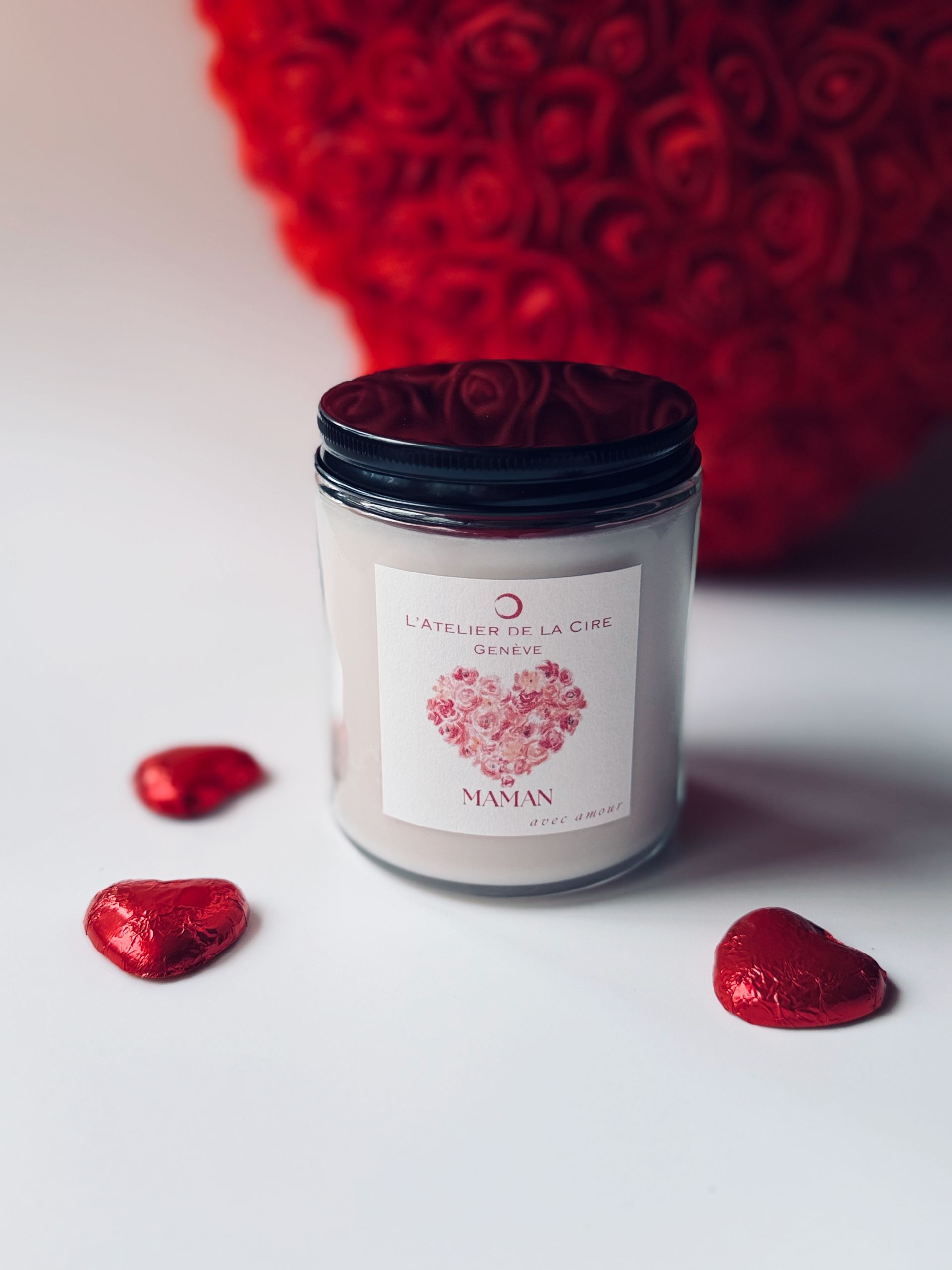 Bougie blanche ornée d'un cœur rose et de pétales de rose, devant des roses rouges sur fond clair.