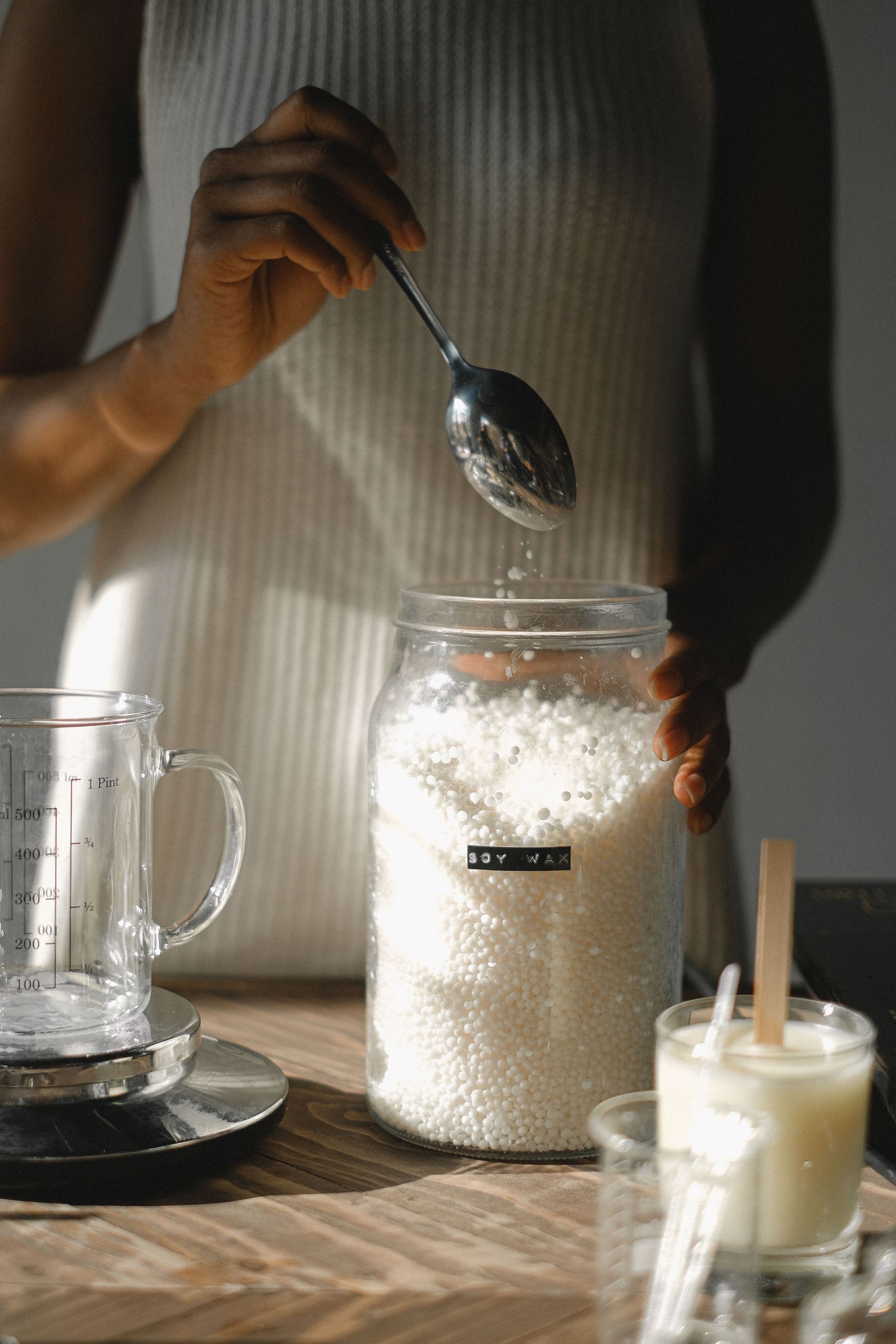 Une personne en pull blanc verse du sucre à la cuillère dans un bocal en verre, à côté de tasses à café posées sur une table en bois.