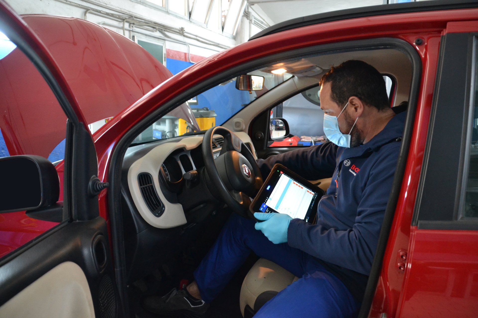 Un meccanico a bordo di un'auto rossa usa un tablet mentre è seduto al posto di guida, con il cofano aperto, in un'officina.