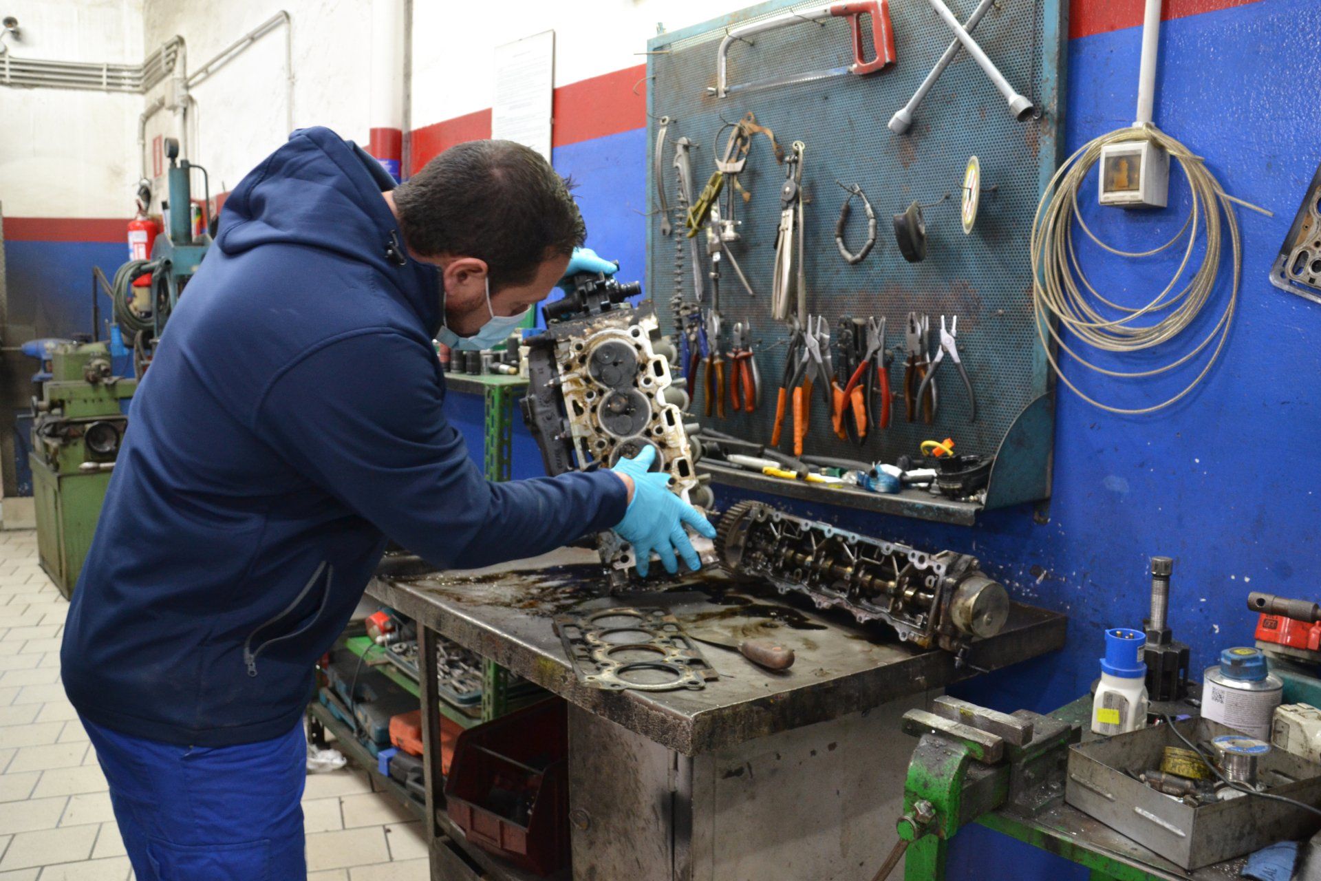 Un meccanico con una felpa blu lavora su un motore in un'officina, con gli attrezzi appesi al muro.
