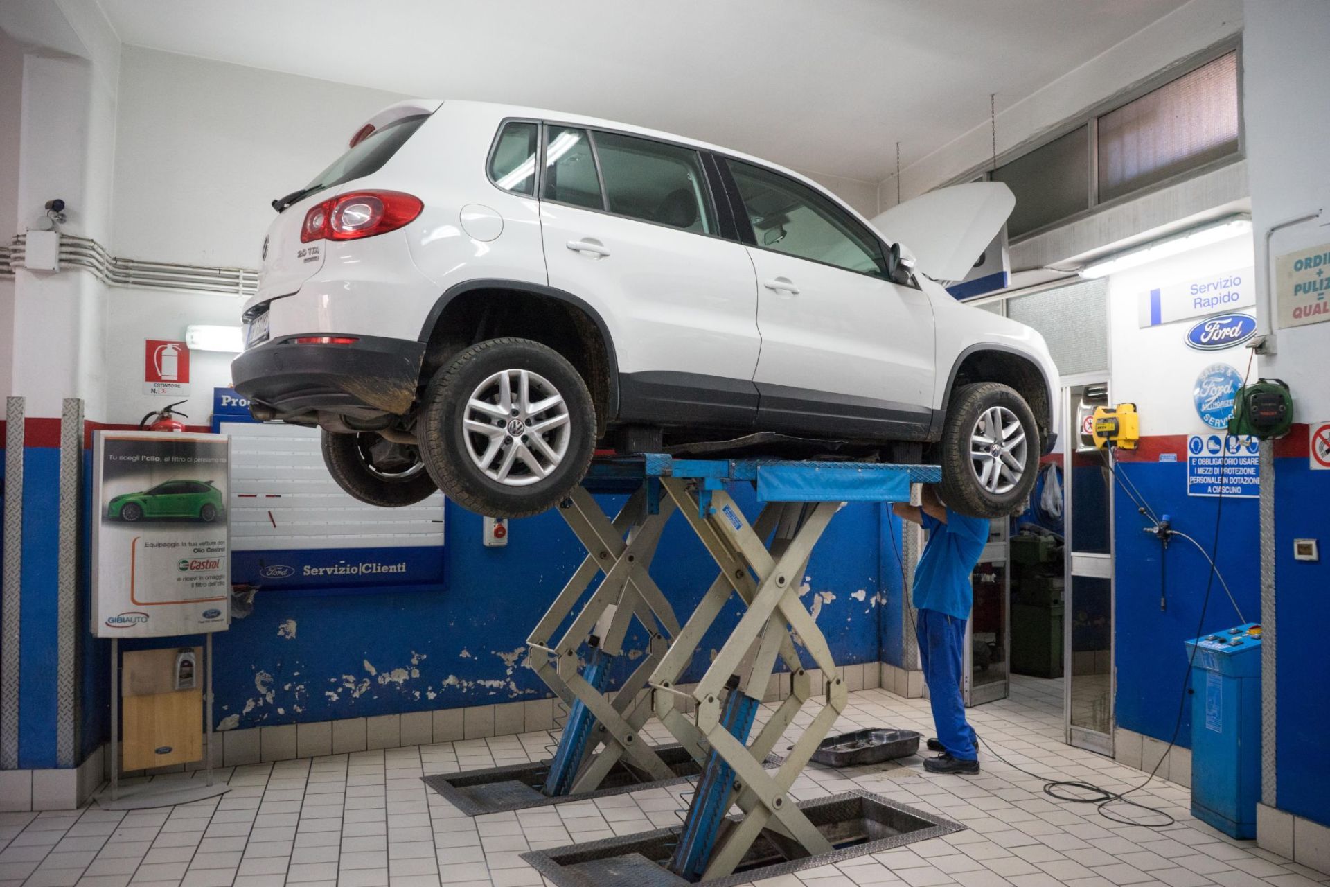 Un SUV bianco è sollevato su un ponte sollevatore in un'officina meccanica, con un meccanico che lavora al di sotto.