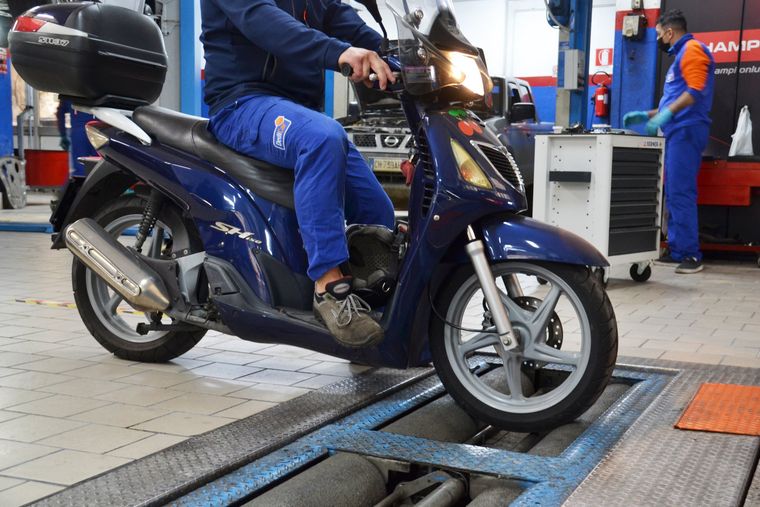 Uno scooter blu su una rampa di ispezione veicoli in un'officina, con un conducente seduto e del personale nelle vicinanze.