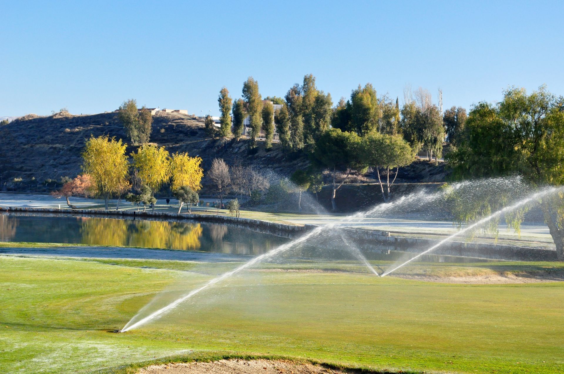A sprinkler sprays water across a bright green golf course near a calm lake and autumn-colored trees under a blue sky.