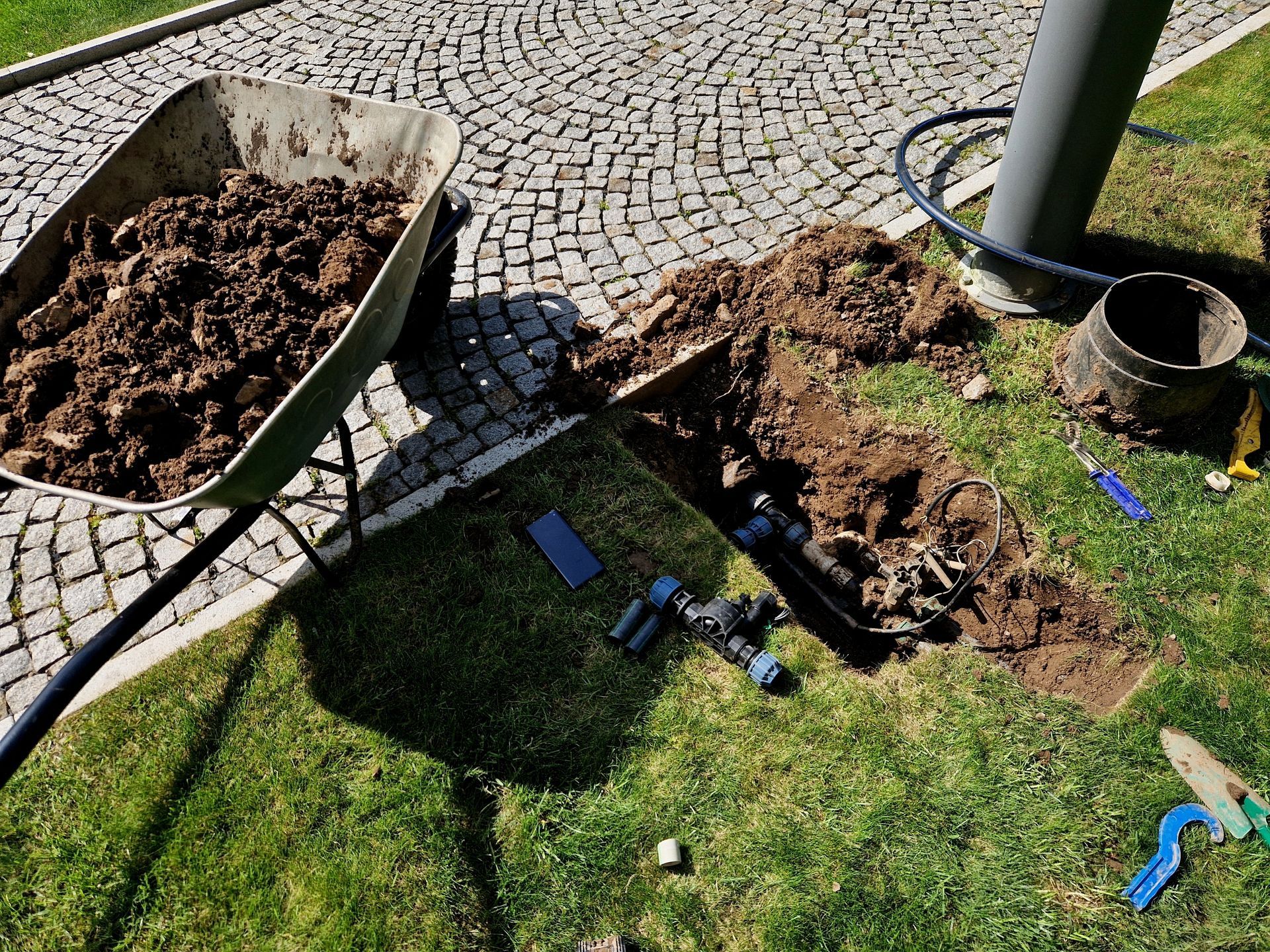 A wheelbarrow full of dirt sits next to an open trench in a grassy area, revealing exposed irrigation pipes and tools.