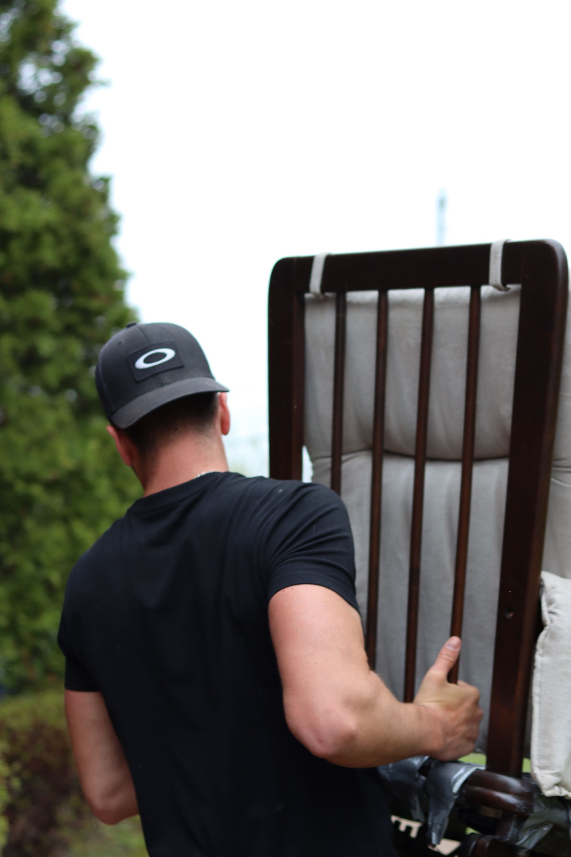 Une personne en chemise noire transporte une chaise à l'extérieur, près d'un arbre et d'une clôture.