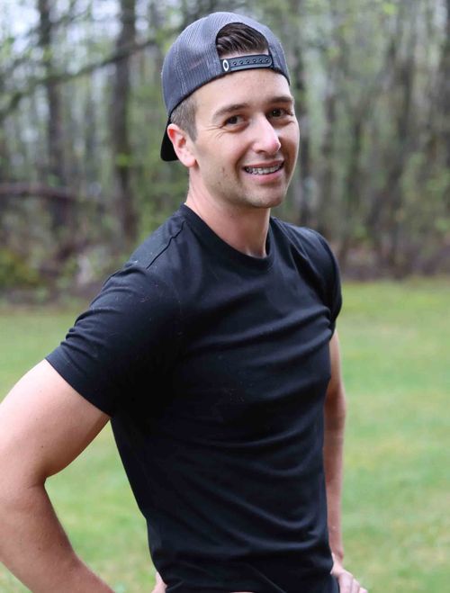 Un homme souriant, vêtu d'un t-shirt noir et d'une casquette à l'envers, se tient debout à l'extérieur, sur l'herbe.
