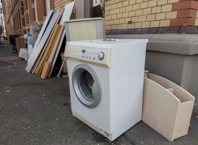 Machine à laver blanche abandonnée et cartons près d'un bâtiment en briques, avec des planches empilées à proximité.