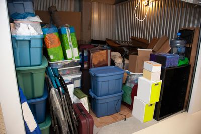 Pièce de stockage encombrée de bacs en plastique, de boîtes et d'objets pliés empilés le long d'un mur ondulé.