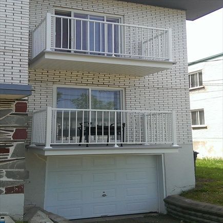 Bâtiment de deux étages en briques blanches. Balcon à l'étage et balcon au rez-de-chaussée. Rambardes blanches. Porte de garage blanche au niveau du sol.