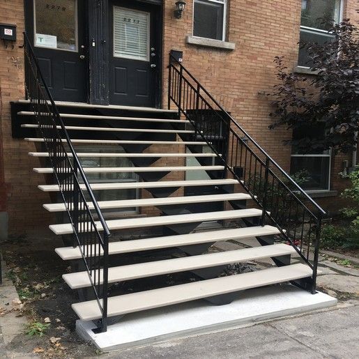 Un escalier extérieur mène aux doubles portes noires d'un bâtiment, avec des rampes noires et des marches blanc cassé.