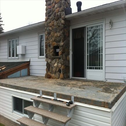 Une maison avec une cheminée en pierre, une porte-fenêtre coulissante et une terrasse en béton avec des marches.