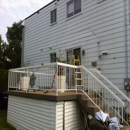 Vue arrière d'une maison blanche à deux étages avec terrasse et escalier. Une échelle jaune se trouve près de la porte coulissante.