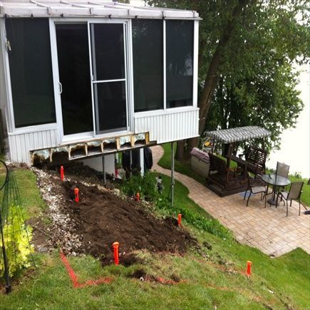 Maison avec terrasse en construction sur une colline herbeuse, surplombant un lac. Des piquets délimitent le terrain.