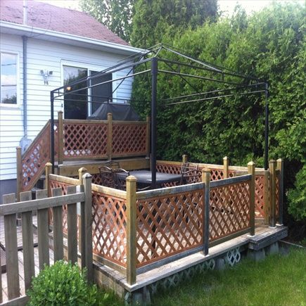 Terrasse en bois avec treillis, pergola en métal foncé et balustrade en treillis, jouxtant une maison et de la verdure.