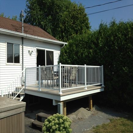 Maison blanche avec terrasse surélevée et balustrade blanche. Des buissons verts et un sol gravillonné entourent la structure.