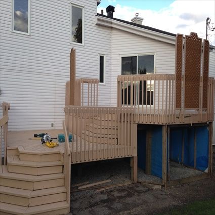 Terrasse en bois avec escalier adossée à une maison blanche. Outils sur la terrasse, bâche bleue sous la structure.