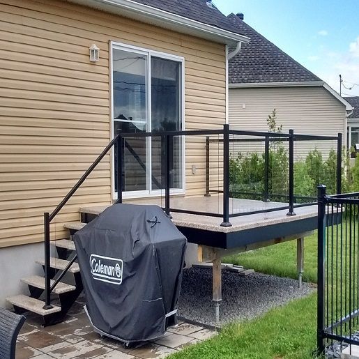 Terrasse avec rambarde noire, escalier et barbecue couvert, jouxtant une maison au bardage beige et à la porte-fenêtre coulissante.