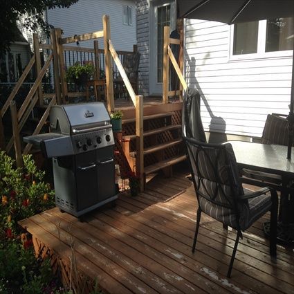 Une terrasse dans le jardin avec barbecue, escalier et table avec chaises, à l'ombre partielle.