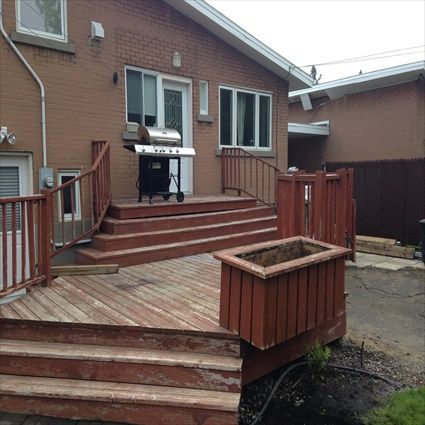 Une terrasse avec escalier, rambarde et barbecue, adossée à une maison en briques. Une jardinière se trouve au premier plan.