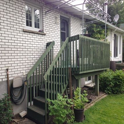 Terrasse en bois vert avec escalier attenant à une maison en briques blanches. Un tuyau d'arrosage est accroché au mur.
