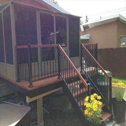 Terrasse couverte avec escalier et rambarde noire ; fleurs jaunes à l’avant. Couleurs marron et noir. Aménagement extérieur.