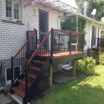 Une terrasse arrière avec escalier, balustrade noire et finitions en bois brun-rouge. Une maison en briques blanches se dresse à l'arrière-plan.
