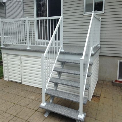 Un escalier extérieur à rampe blanche mène à une terrasse avec rangements. Revêtement extérieur gris et terrasse en béton.