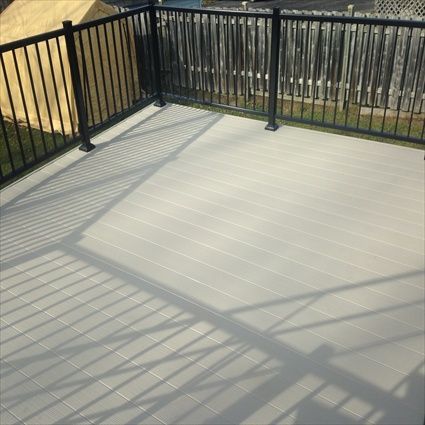 Une terrasse en composite gris clair avec une rambarde en métal noir, projetant des ombres au soleil. Aménagement d'un jardin.