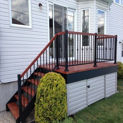 Terrasse avec rambarde noire, marches marron et espace de rangement sous une maison au bardage blanc.