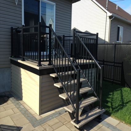 Rambarde et escalier noirs menant à une terrasse attenante à une maison au bardage beige et au patio en briques.