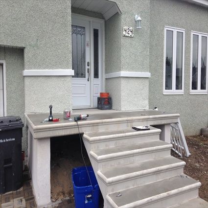 Des marches et un porche en béton gris mènent à une porte d'entrée blanche d'une maison au crépi vert clair.
