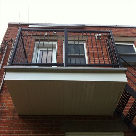 Balcon en métal noir avec rambarde, fixé à un bâtiment en briques, au-dessus d'un plafond à panneaux blancs.