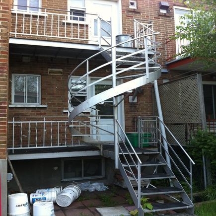 Vue extérieure d'un immeuble avec un escalier en colimaçon argenté menant à un balcon au deuxième étage. Des pots de peinture sont posés en dessous.