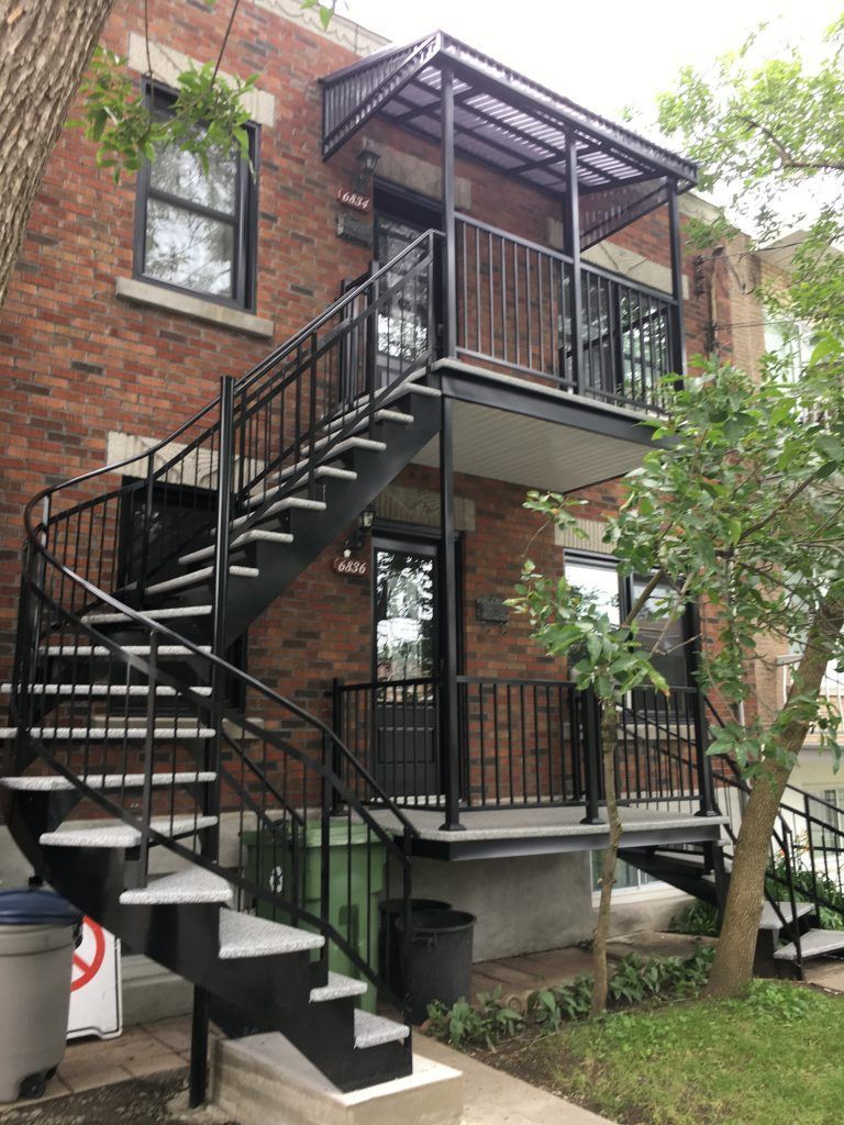 Bâtiment en briques avec un escalier extérieur en métal noir menant à un balcon avec une entrée couverte.