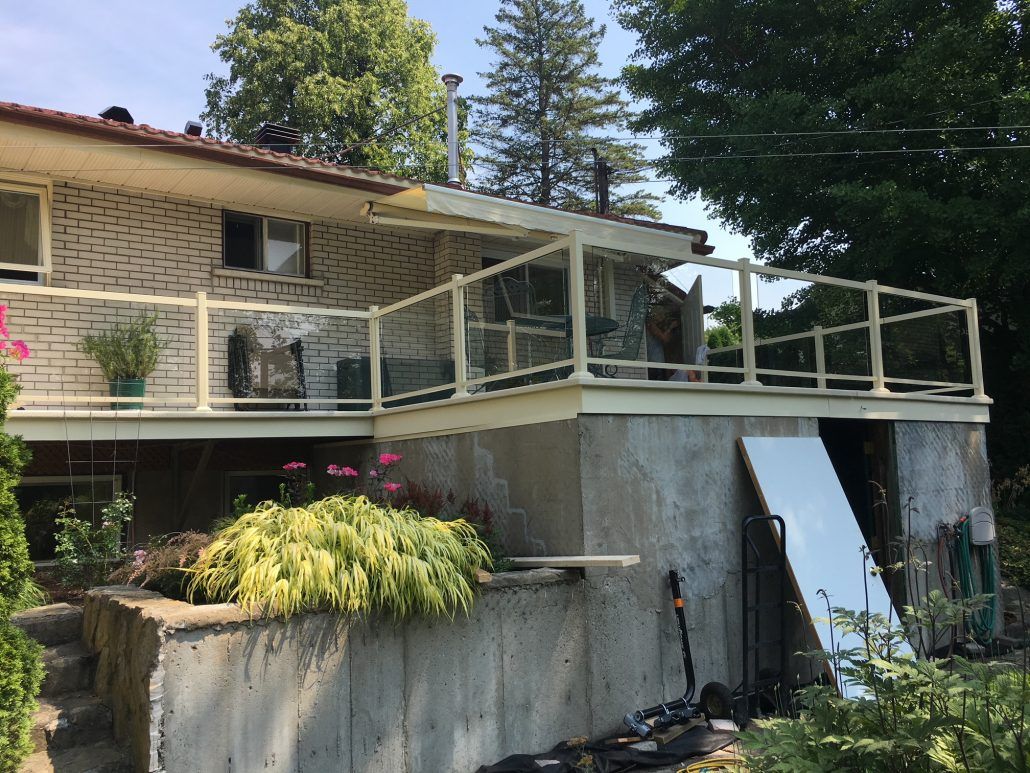 Terrasse beige avec panneaux de verre sur fondation en béton, attenante à une maison en briques claires.