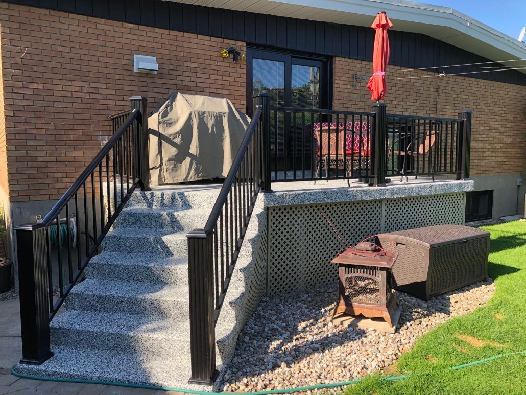 Terrasse arrière avec escalier, rambarde noire, barbecue couvert et parasol rouge.