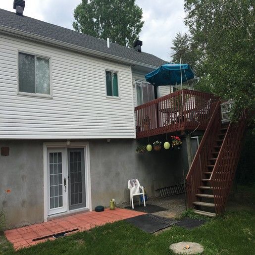 Maison à deux étages avec une terrasse en bois, un escalier et une porte donnant sur un patio carrelé de rouge.