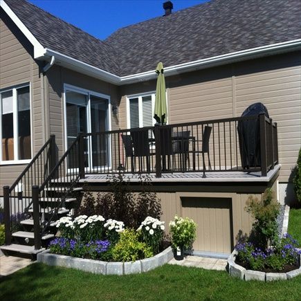 Terrasse arrière avec rambarde foncée, escalier et table. Fleurs dans le jardin. Maison beige avec un parasol vert.