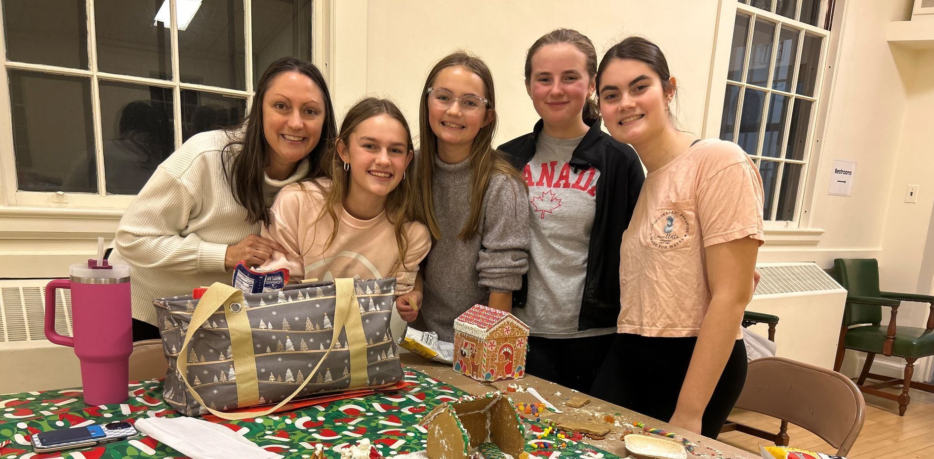 One woman and four girls standing next to their gingerbread house on a table.