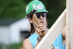 A woman wearing a hard hat and sunglasses is holding a hammer and a piece of wood.