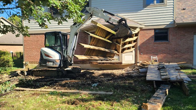 Collapsed deck and stairs beside a house, with a small excavator and scattered debris in the yard.